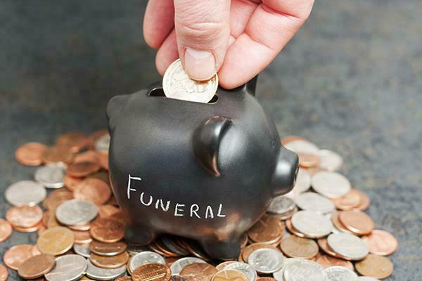 A coin piggy-bank with "Funeral" written on it sitting on a pile of coins with a hand dropping a gold coin into the slot, stock image used by Beth El Mausoleum for funeral costs page.