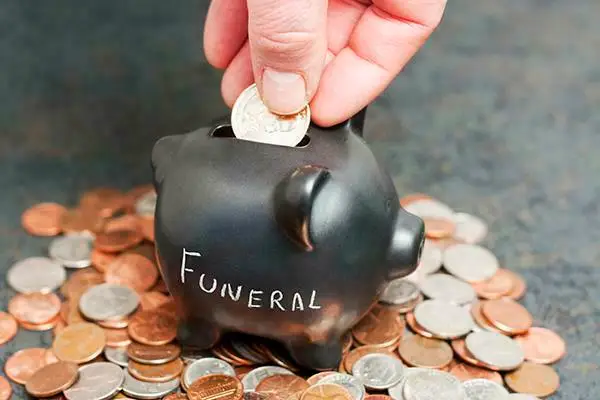 A coin piggy-bank with "Funeral" written on it sitting on a pile of coins with a hand dropping a gold coin into the slot, stock image used by Beth El Mausoleum for funeral costs page.